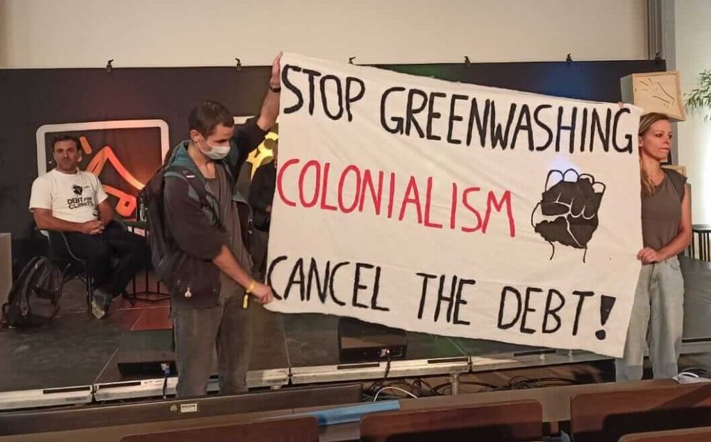 Two people holding a banner in the front of a lecture hall. The banner reads "STOP GREENWASHING COLONIALISM - CANCEL THE DEBT!" and a fist.